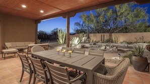 Un patio al aire libre con techo de madera, una mesa de comedor con sillas, un sofá y diversas plantas del desierto.