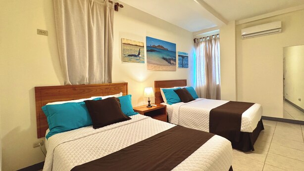 A hotel room with two beds, each with a wooden headboard, white and brown bedding, and a nightstand with a lamp. There are wall-mounted air conditioning units and three framed pictures depicting ocean scenes. - This two-bedroom is a perfect island getaway. (Puerto Ayora, Santa Cruz Island)