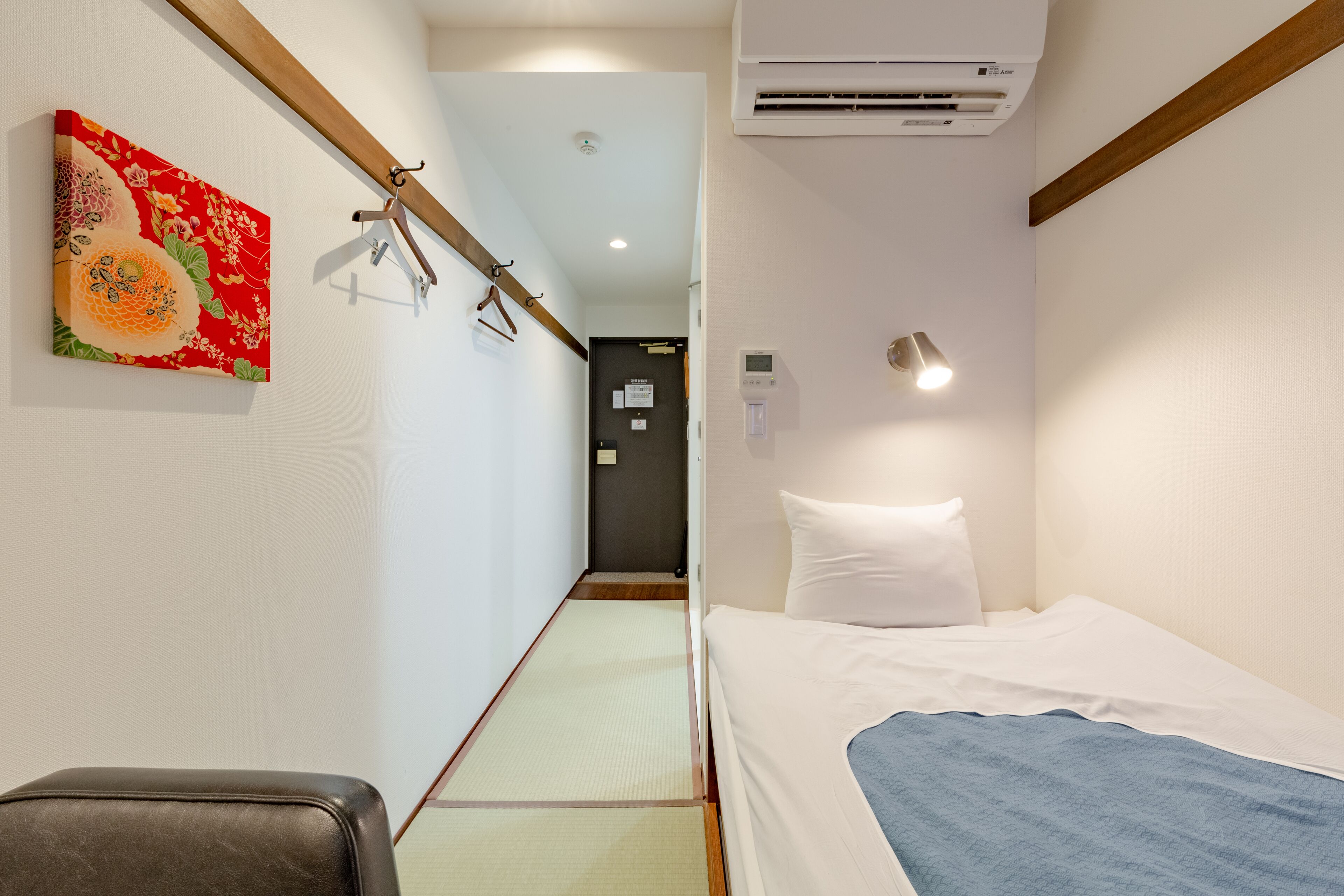 A hotel room with a bed, a wall-mounted air conditioner, a painting, and a hallway leading to another room.