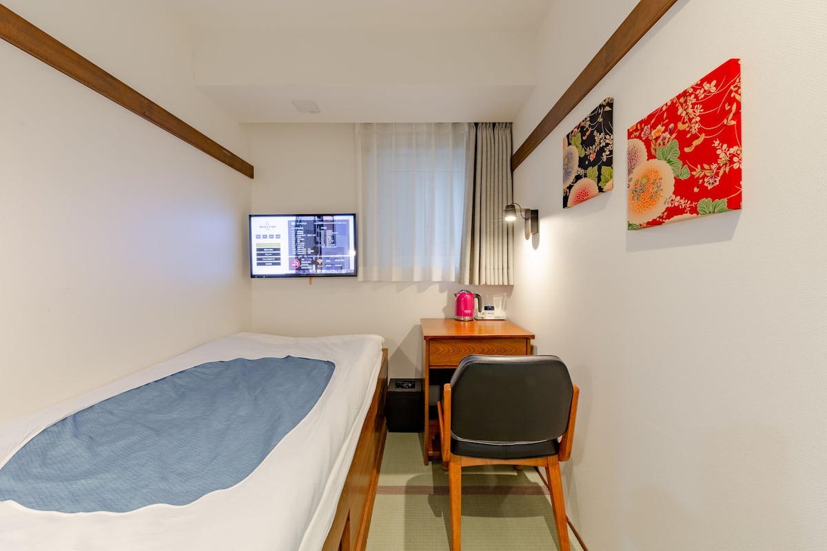 A hotel room with a single bed, a desk with a chair, a flat-screen TV, and two decorative wall hangings.