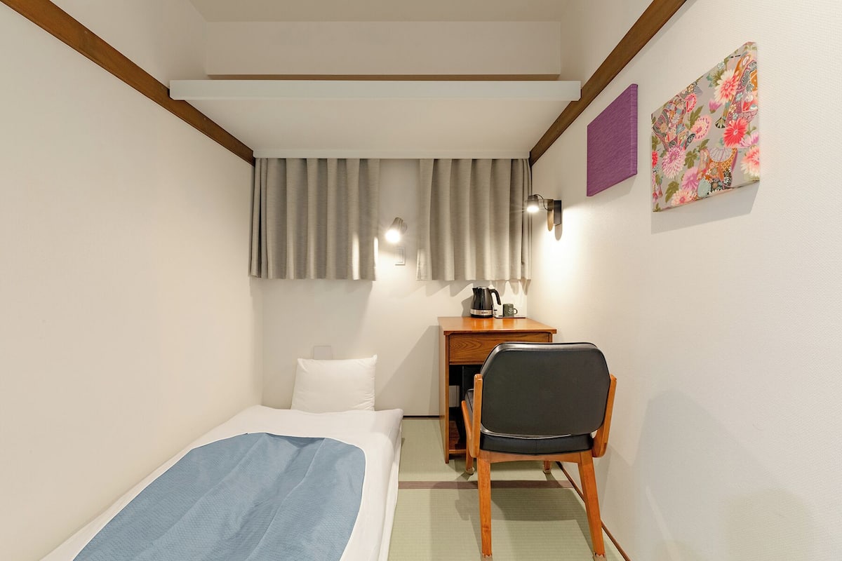 A hotel room with a bed, a desk with a chair, a nightstand, a lamp, and a painting on the wall.