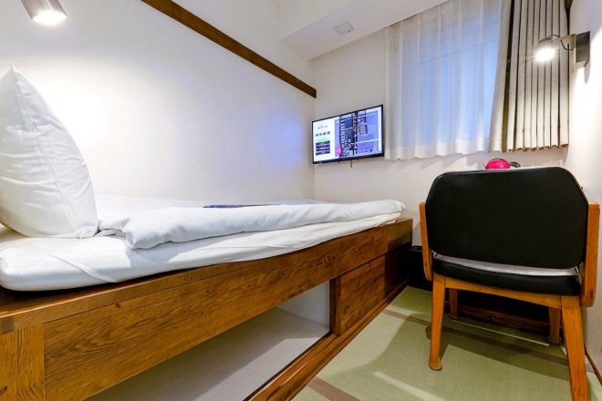 A room with a bed, a chair, a TV mounted on the wall, and a window with blinds.