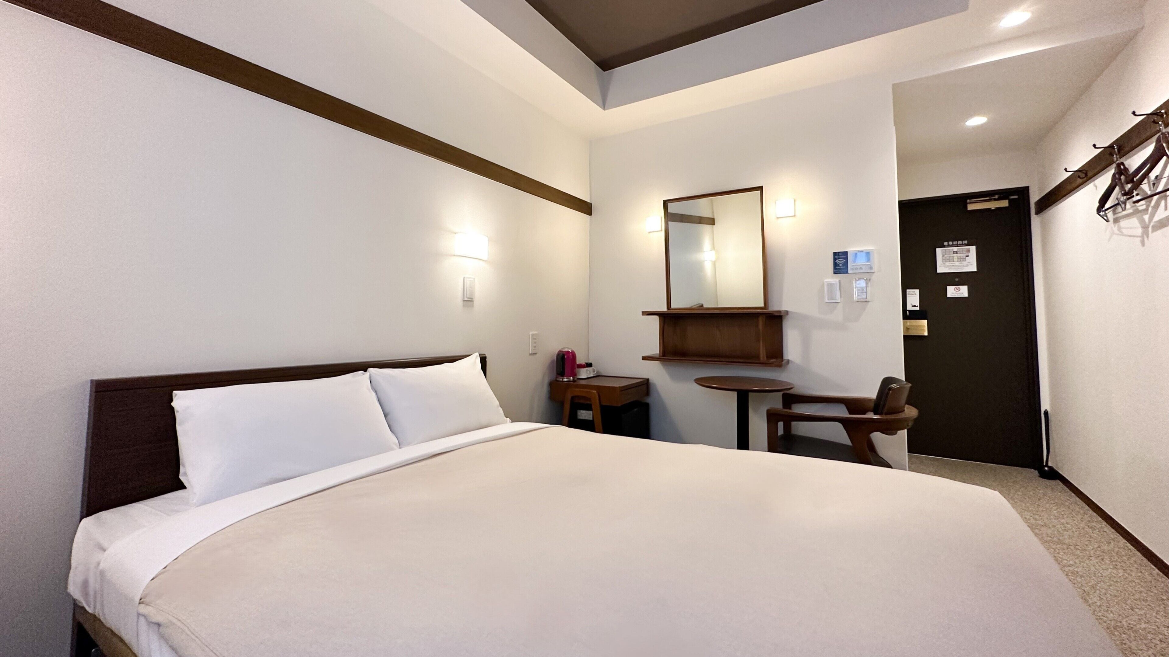 A hotel room with a bed, a desk, a chair, a small table, a mirror, and a door.