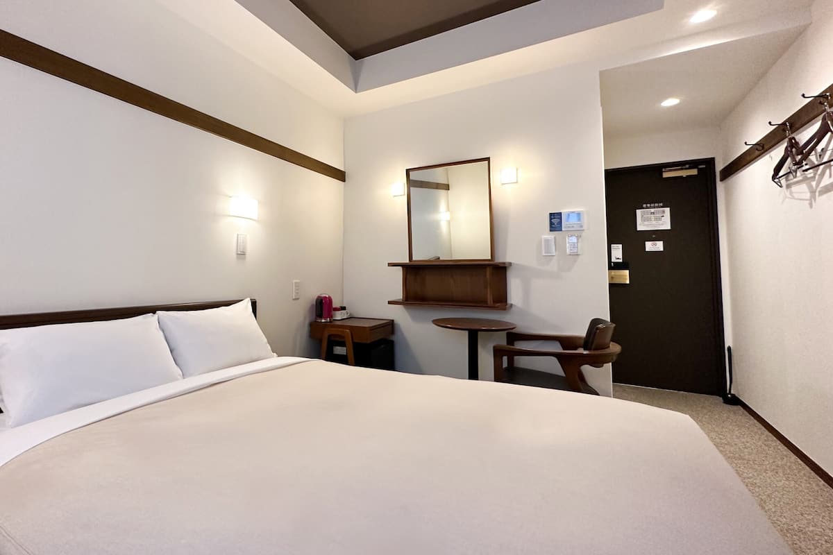 A hotel room with a bed, a desk, a chair, a small table, a mirror, and a door.