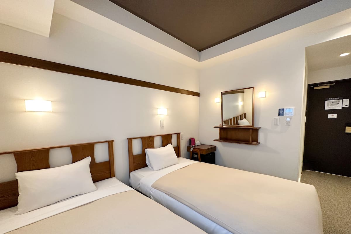 A hotel room with two beds, a wooden headboard, a small bedside table with a lamp, a mirror, and a door with a sign.