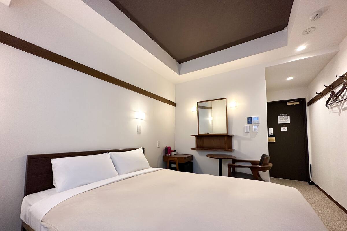 A hotel room with a bed, a small table, a chair, a mirror, and a door.
