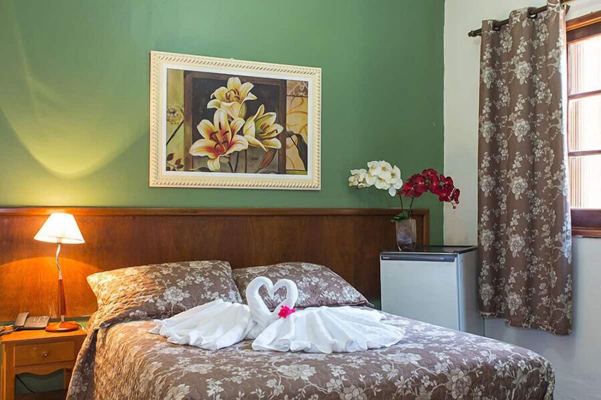 Quarto de hotel com uma cama, uma mesinha de cabeceira com abajur, um pequeno frigorífico, um quadro com flores na parede e uma janela com cortinas.