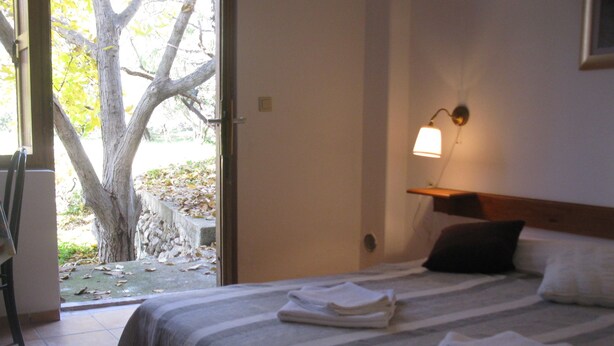A hotel room with a bed, a nightstand, a lamp, and a view of a tree through an open door. - Apartmani Bako Komiza (Komiza)