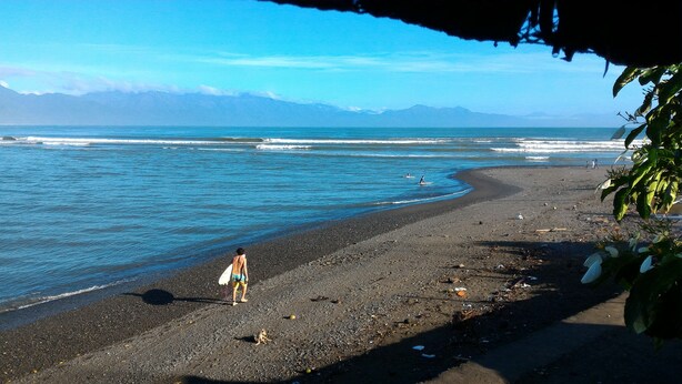 A beach with a rocky shoreline, a person walking with a surfboard, and mountains in the background. - A.S.J. Backpackers Baler (Baler)