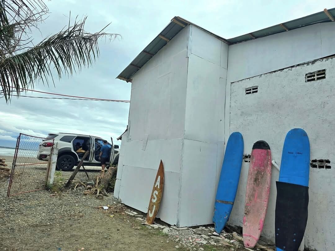 A building with a white exterior, a parked SUV, and three surfboards leaning against the wall.