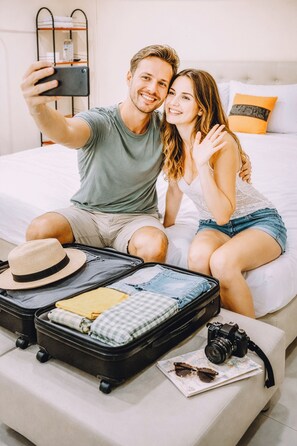 A couple taking a selfie on a bed with a suitcase, camera, and sunglasses on the nightstand. - Khaosan Station Hostel (Bangkok)