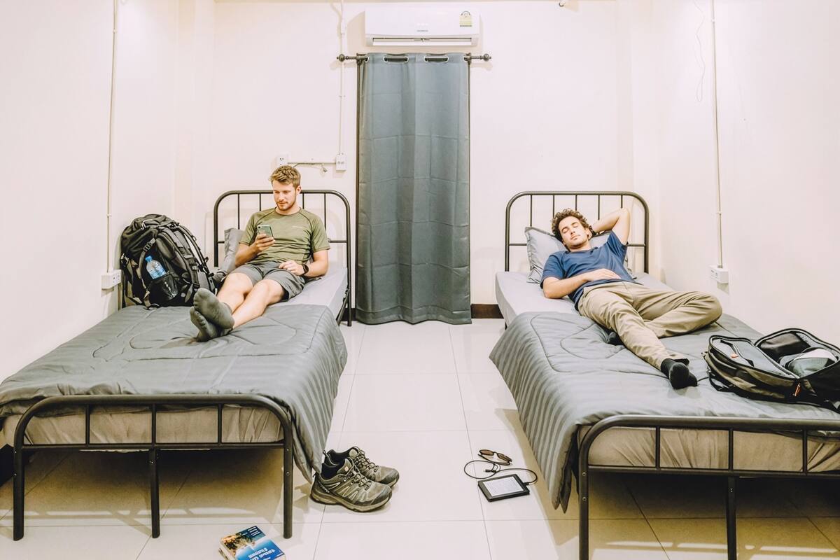 Twee eenpersoonsbedden in een kamer met een gordijn als scheiding, een rugzak op een bed en een paar schoenen op de vloer.