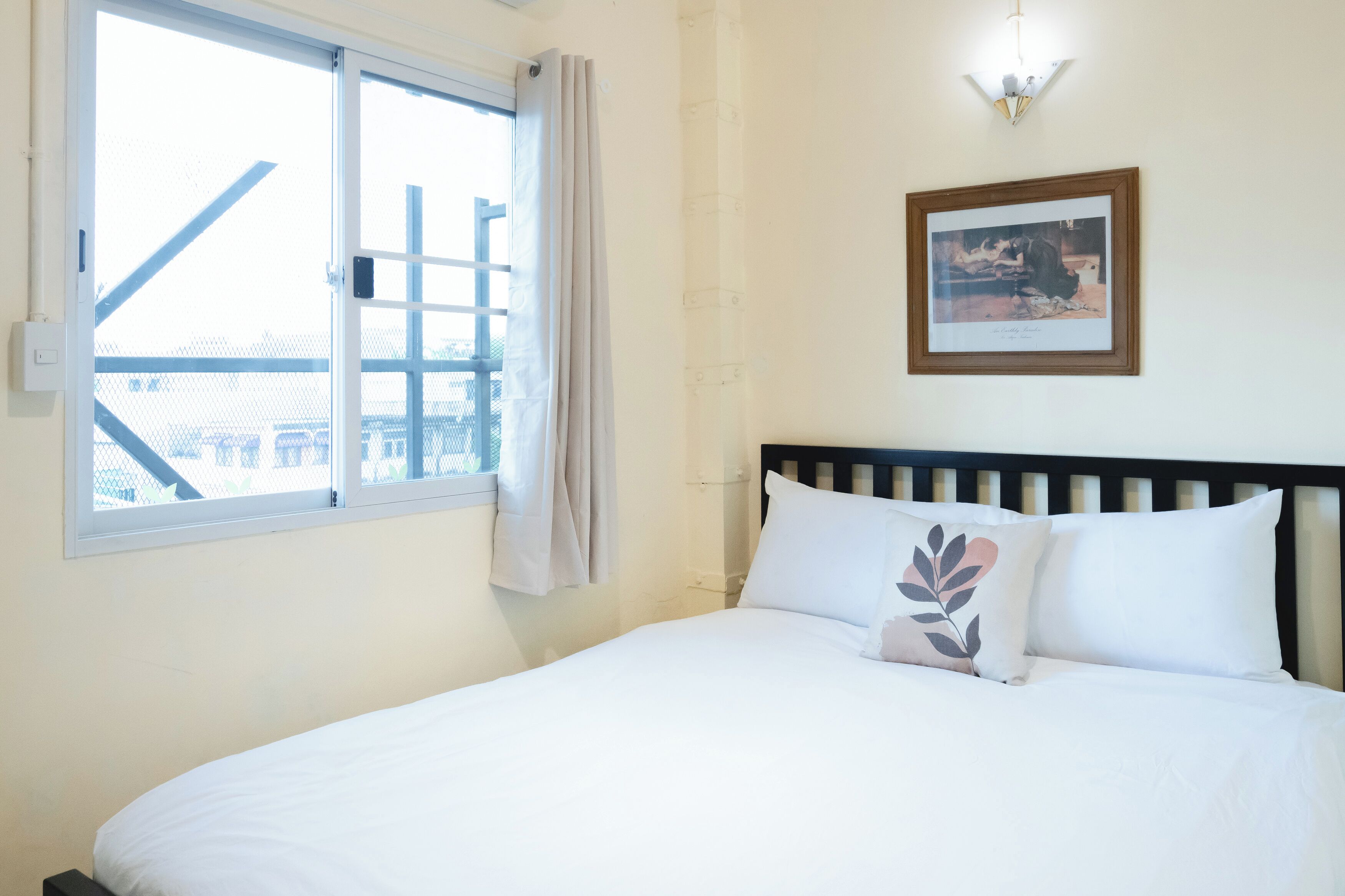Una cama con ropa blanca, una ventana con vistas, una lámpara de pared y un cuadro enmarcado.