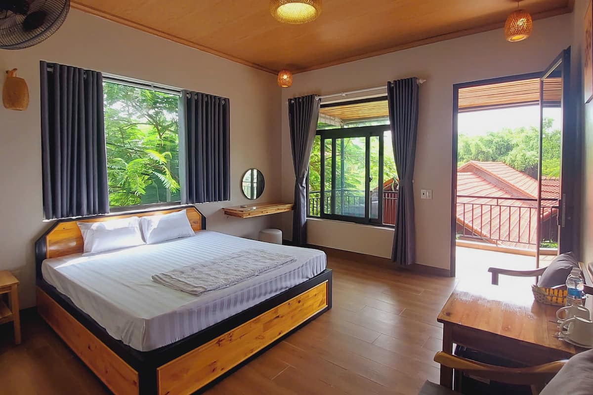 A bedroom with a wooden bed, a nightstand, a desk, and a view of greenery outside.