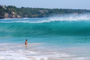 A beach with a large wave, a person in the water, and a coastal area with buildings. - Sunset villa (Ungasan)