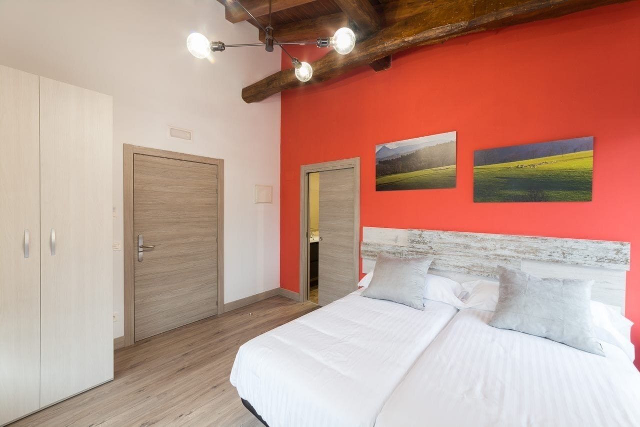 A bedroom with a bed, two doors, and a red wall with two landscape paintings.