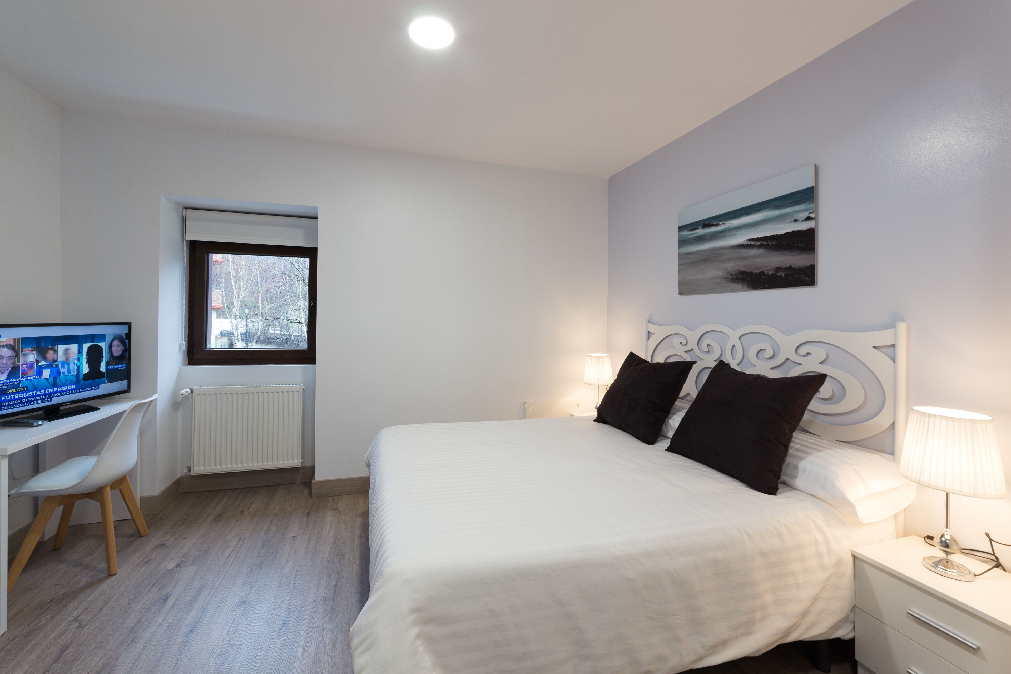 Un dormitorio con cama, escritorio, silla, televisor, ventana y un cuadro en la pared.