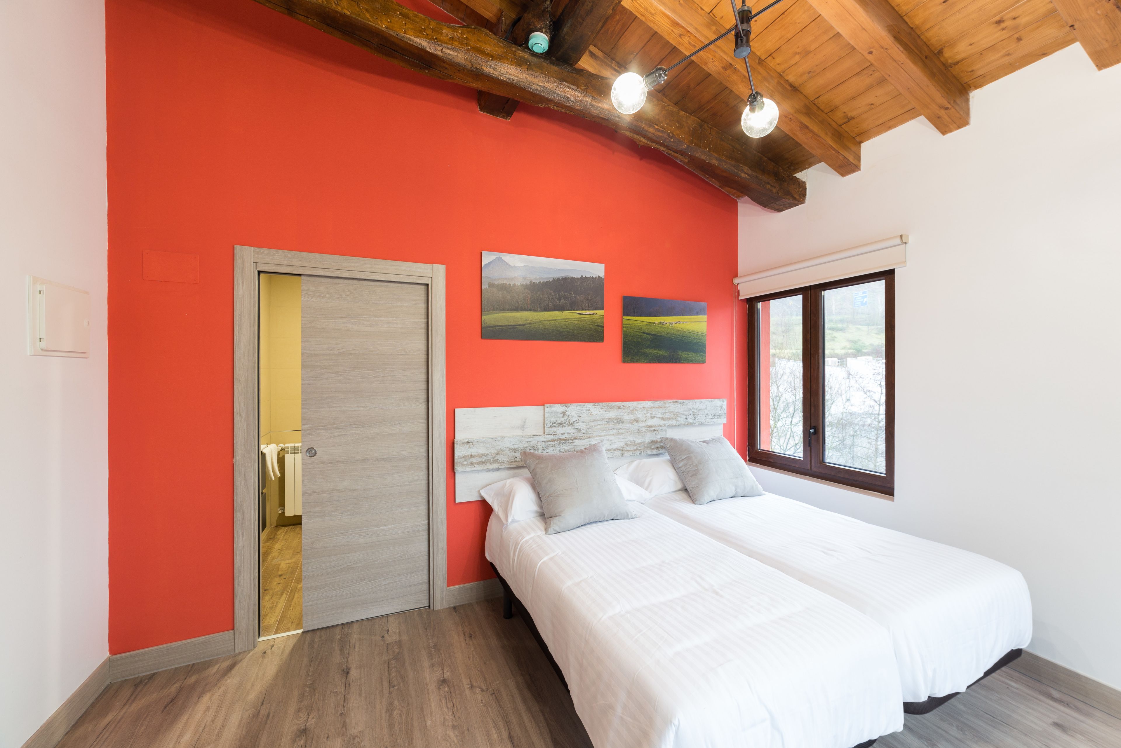 A bedroom with a red wall, wooden ceiling beams, a bed with white bedding, and two landscape paintings.