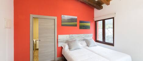 A bedroom with a red wall, wooden ceiling beams, a bed with white bedding, and two landscape paintings.