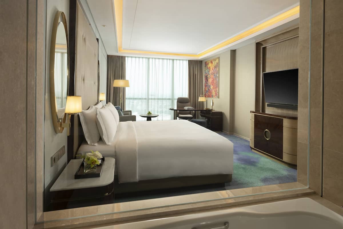 Habitación de hotel con una cama grande, televisión, escritorio con lámpara, una silla y una mesita con flores.