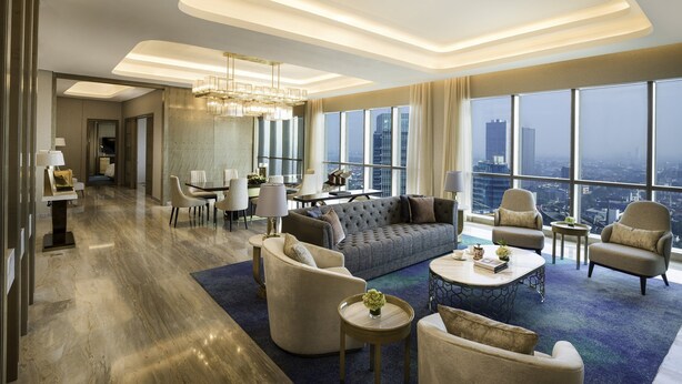A spacious living room with a large sofa, armchairs, and a coffee table. The room has a high ceiling with recessed lighting and large windows offering a city view.