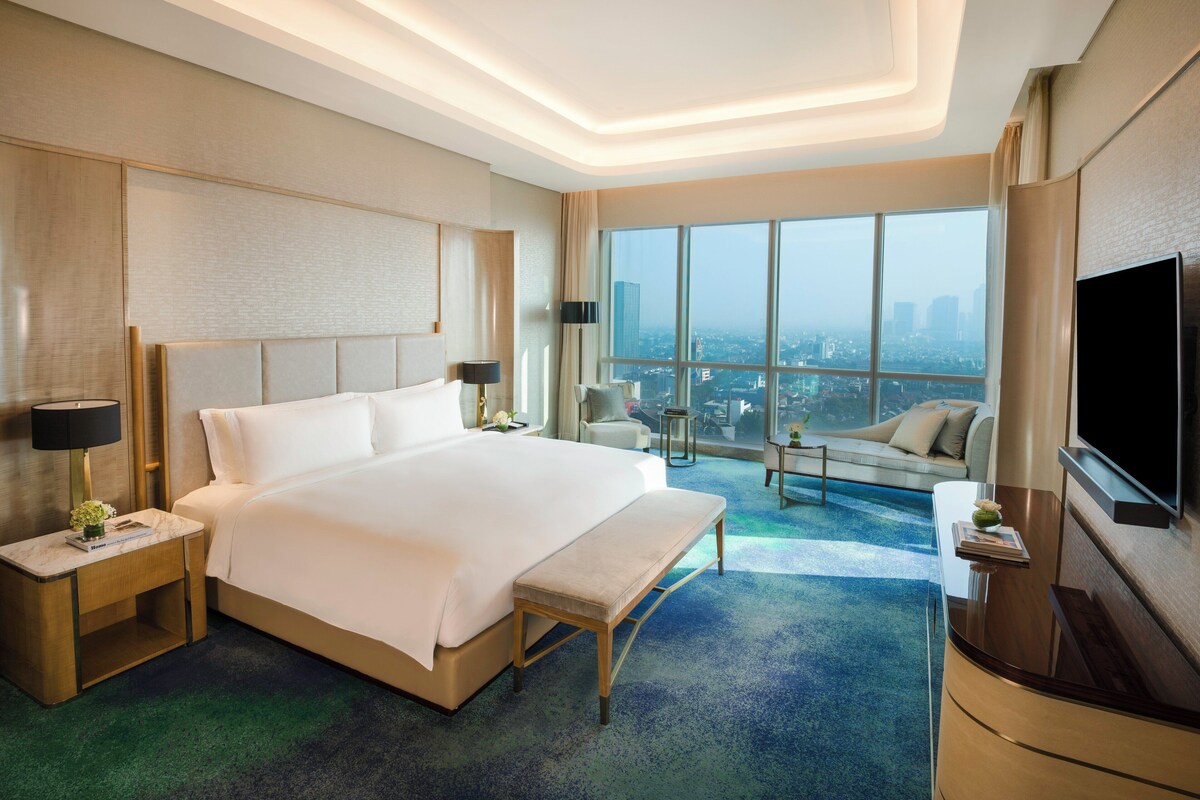 Habitación de hotel moderna con una cama grande, un televisor de pantalla plana y vistas a la ciudad a través de amplios ventanales.