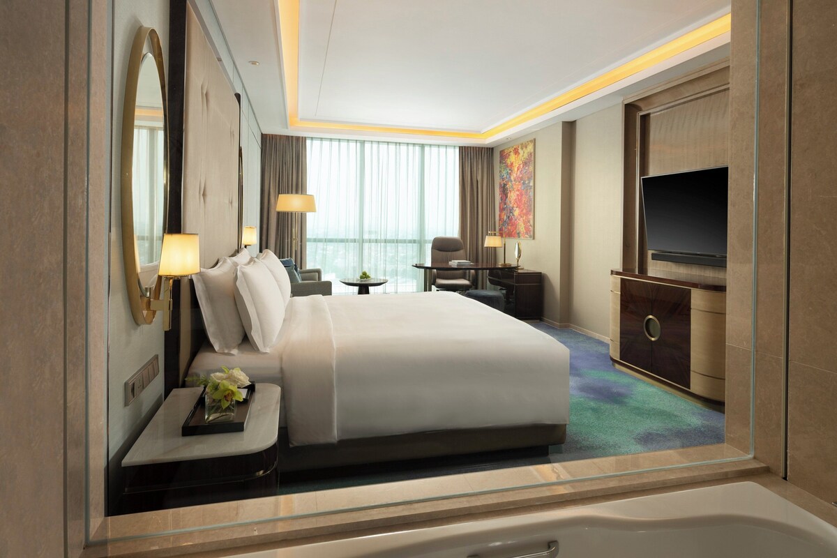 Habitación de hotel con una cama grande, televisión, escritorio con lámpara, una silla y una mesita con flores.