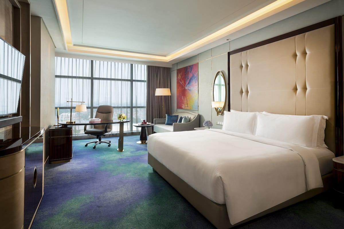 Habitación de hotel con una cama grande, un escritorio con silla, un sofá y un ventanal con cortinas.