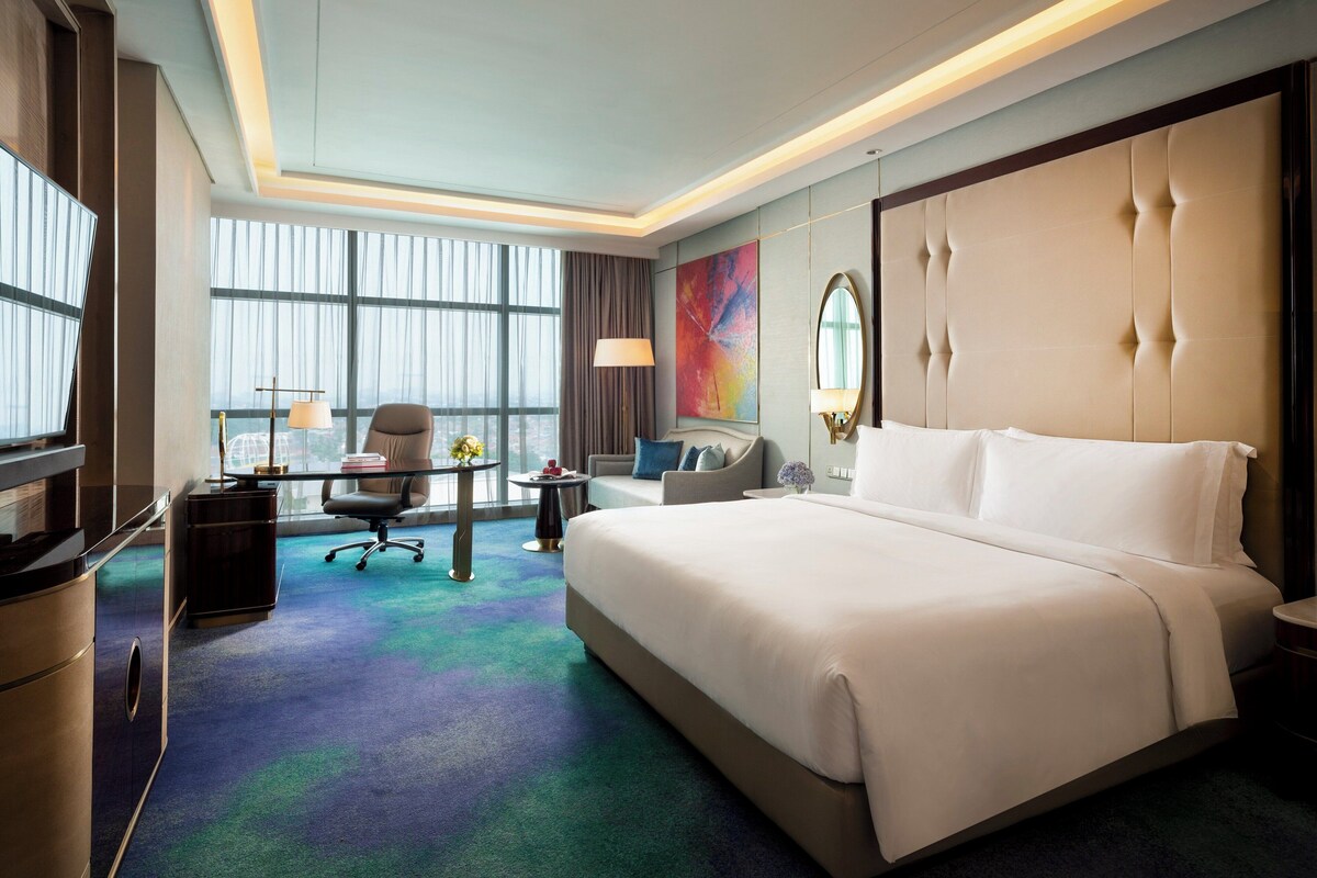 Habitación de hotel con una cama grande, un escritorio con silla, un sofá y un ventanal con cortinas.