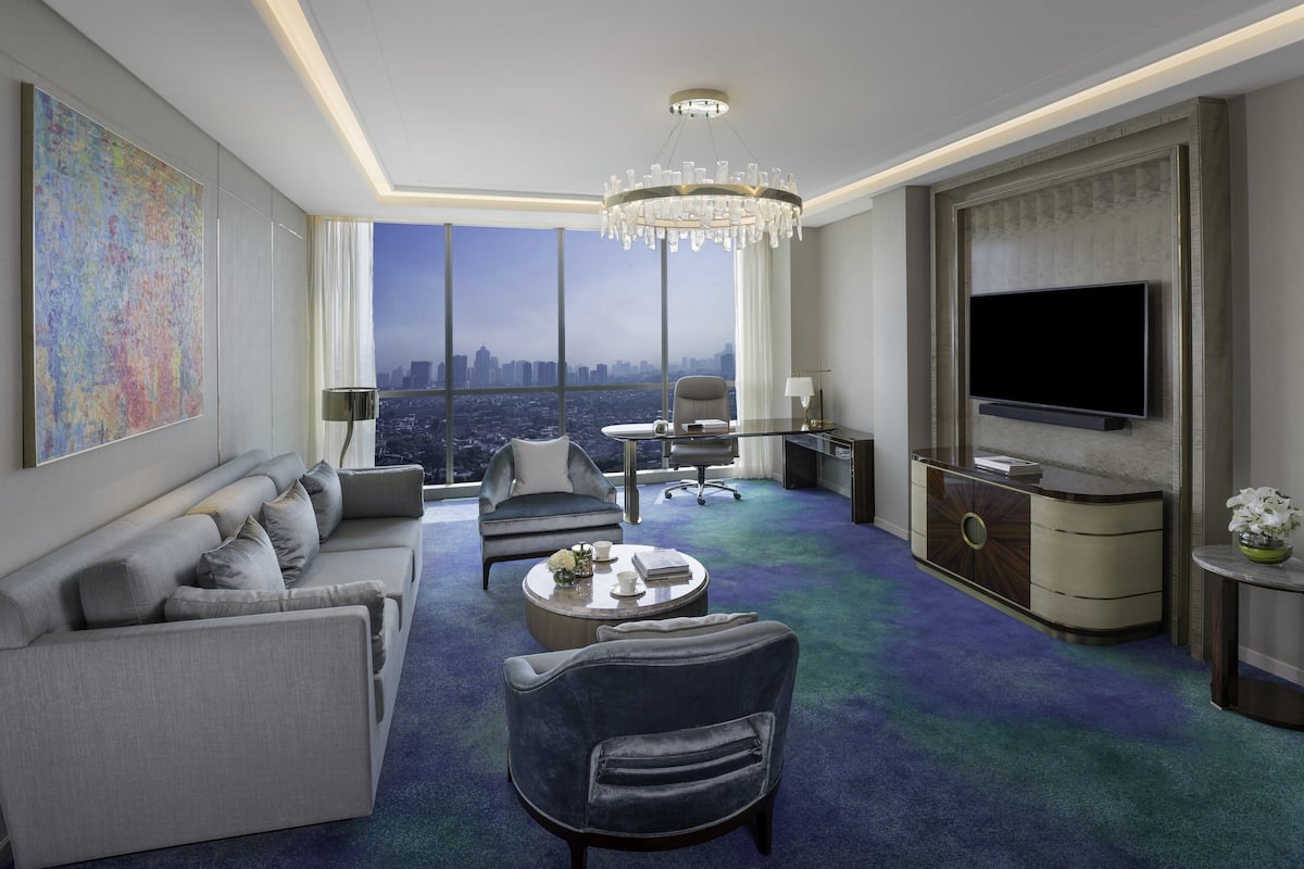 Habitación de hotel moderna con una amplia vista de la ciudad, un candelabro, un televisor de pantalla plana y una zona de estar con sofá y sillones.