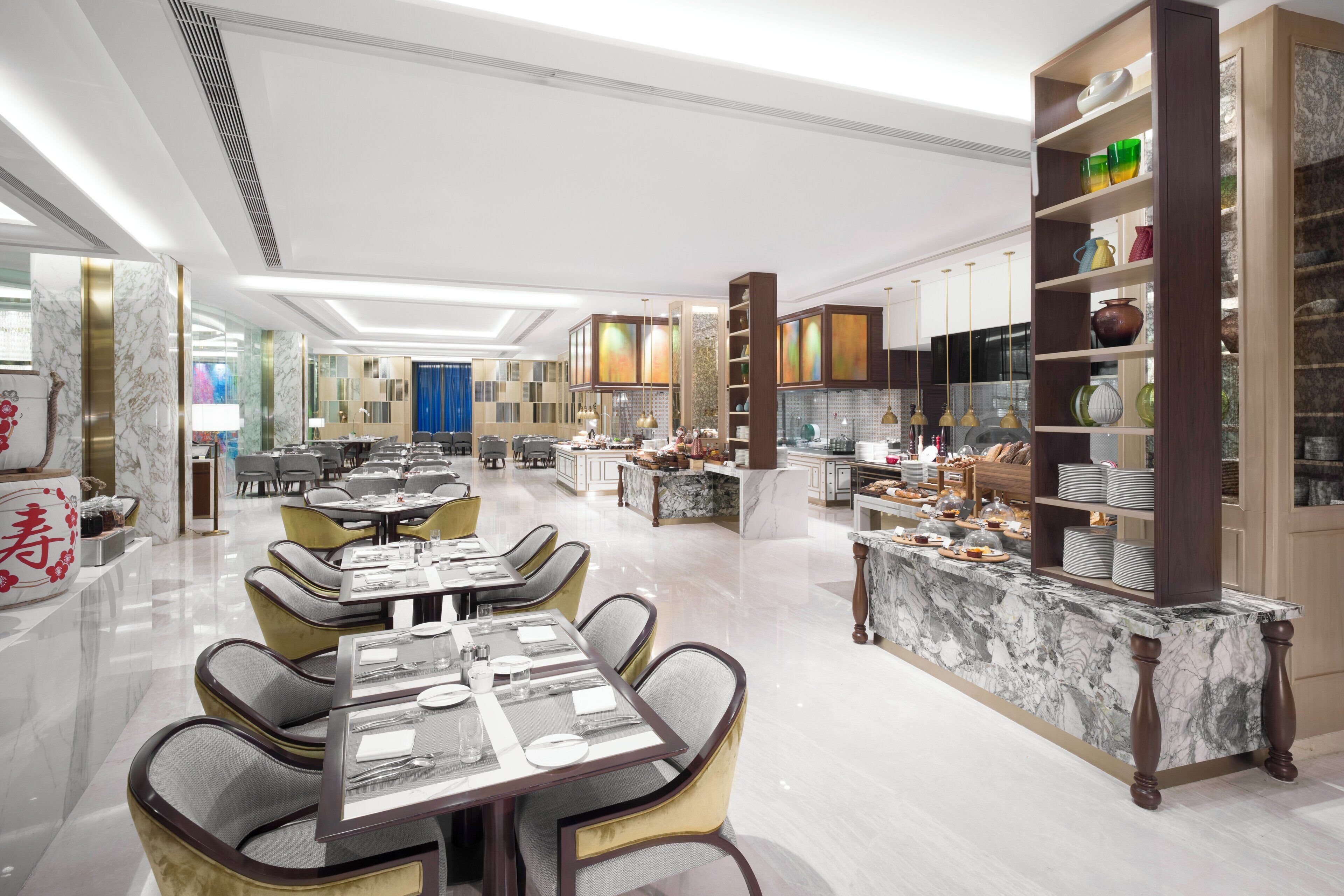 A spacious hotel dining area with multiple tables and chairs, a central island, and a variety of kitchenware displayed on shelves.