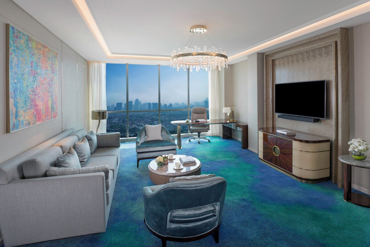 Habitación de hotel moderna con una amplia vista de la ciudad, un candelabro, un televisor de pantalla plana y una zona de estar con sofá y sillones.