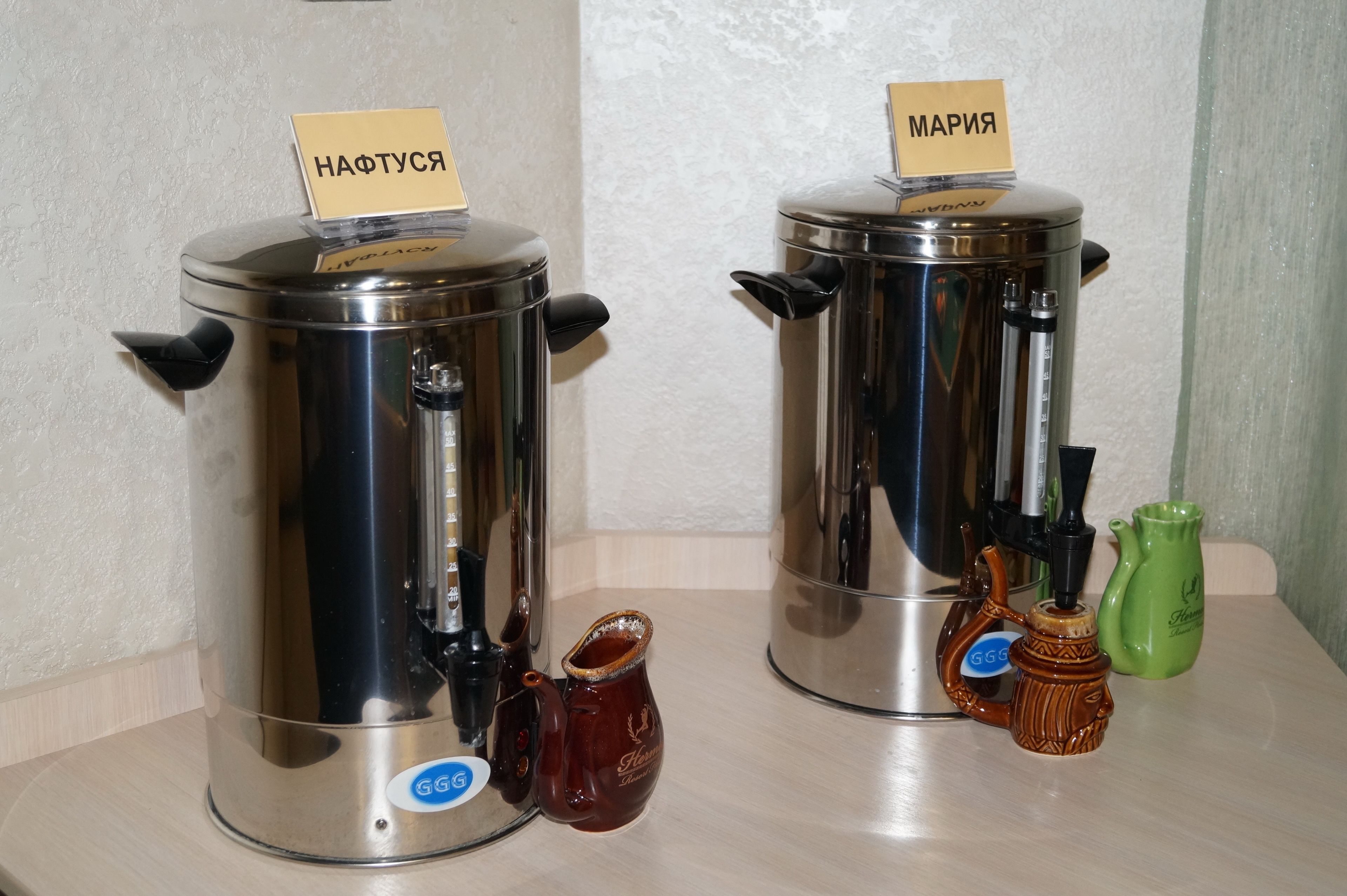 Zwei Edelstahl-Kaffeemaschinen mit Beschriftungen, eine kleine braune Kanne und ein grüner Krug auf einem Tisch.