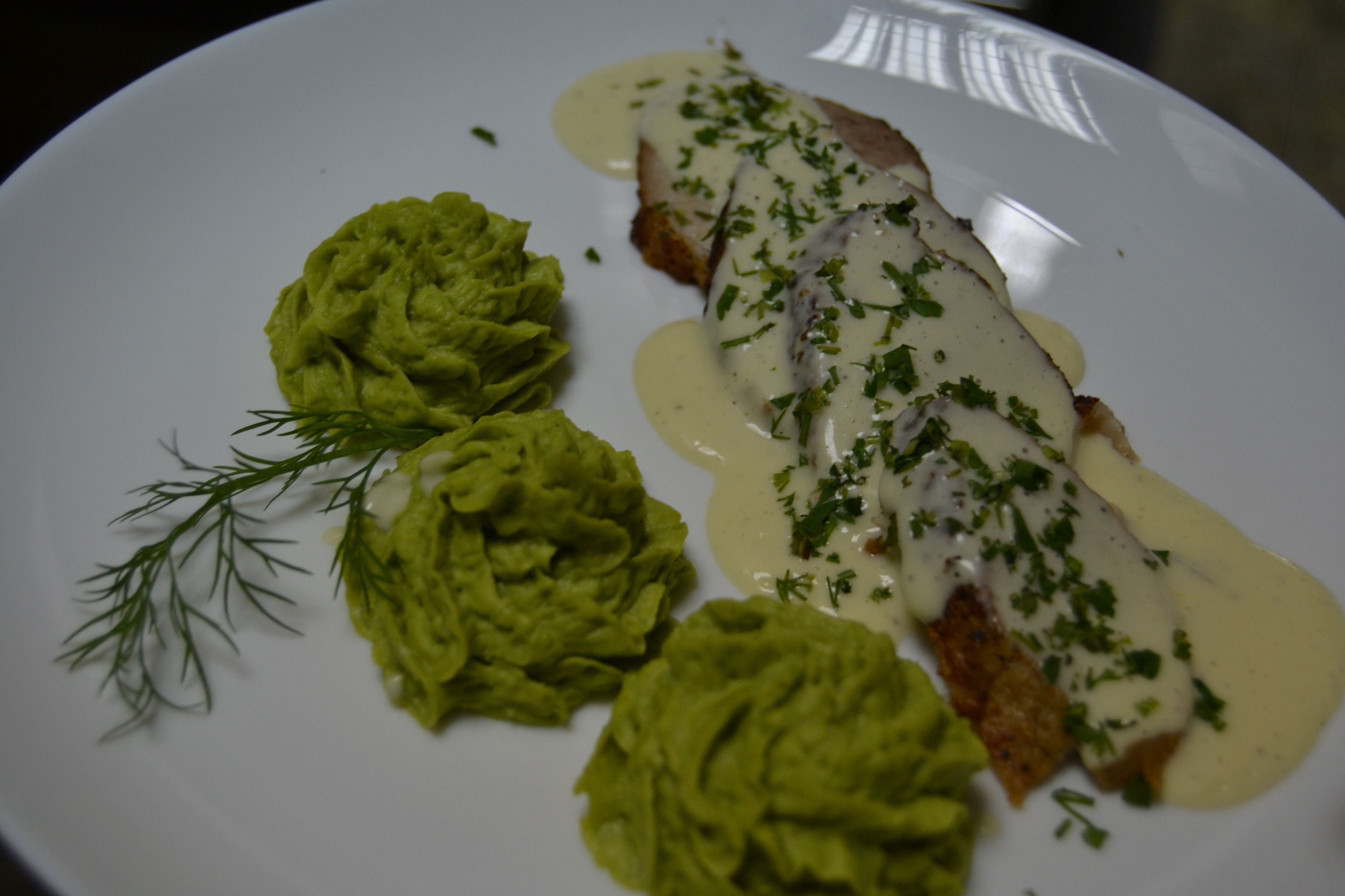 Un plato con un trozo de carne bañado en salsa y adornado con hierbas, acompañado de una cucharada de salsa verde y una ramita de eneldo.