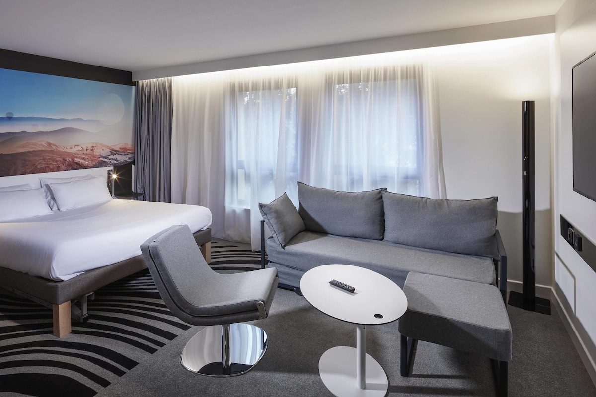 A modern hotel room with a bed, a sofa, a chair, a small table, and a TV.