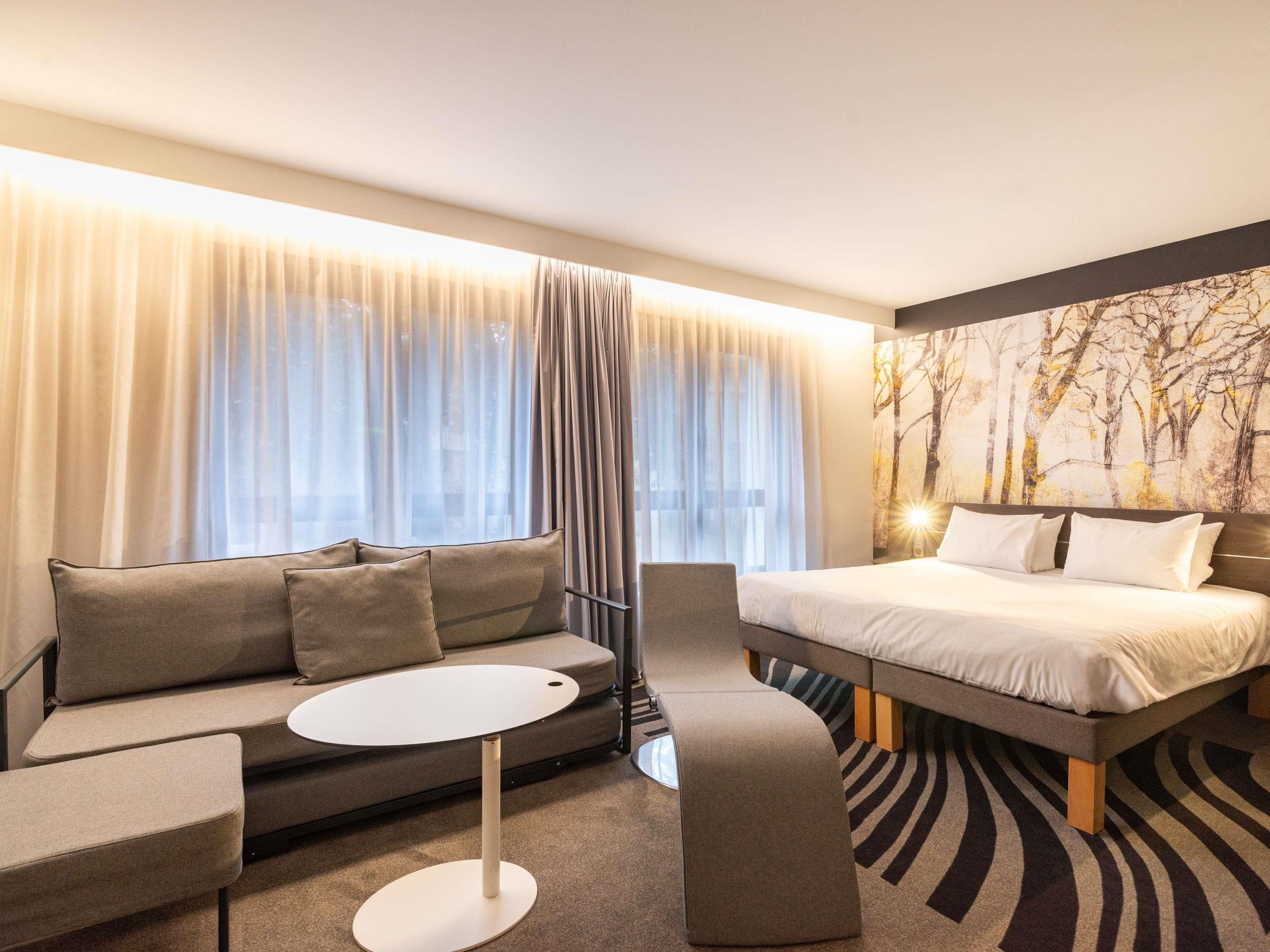Quarto de hotel moderno com sofá, mesa redonda, cama e uma grande janela com cortinas leves.