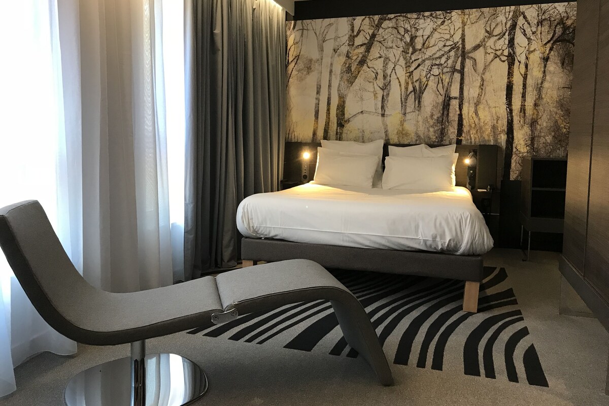 Un dormitorio moderno con una cama, una silla, una mesita de noche y una pared con un mural de un árbol.