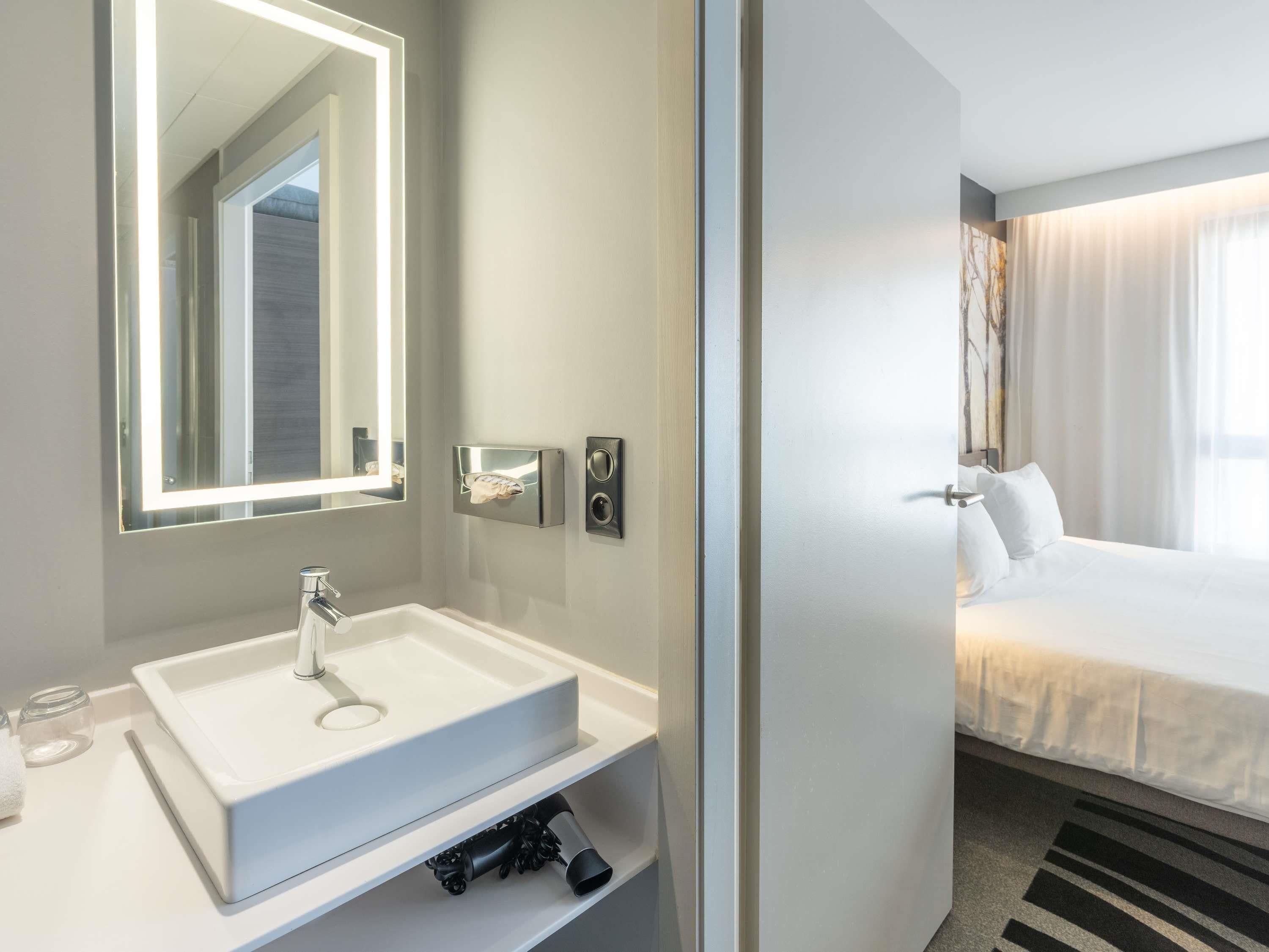 A modern bathroom with a large mirror, a white sink, and a towel dispenser.