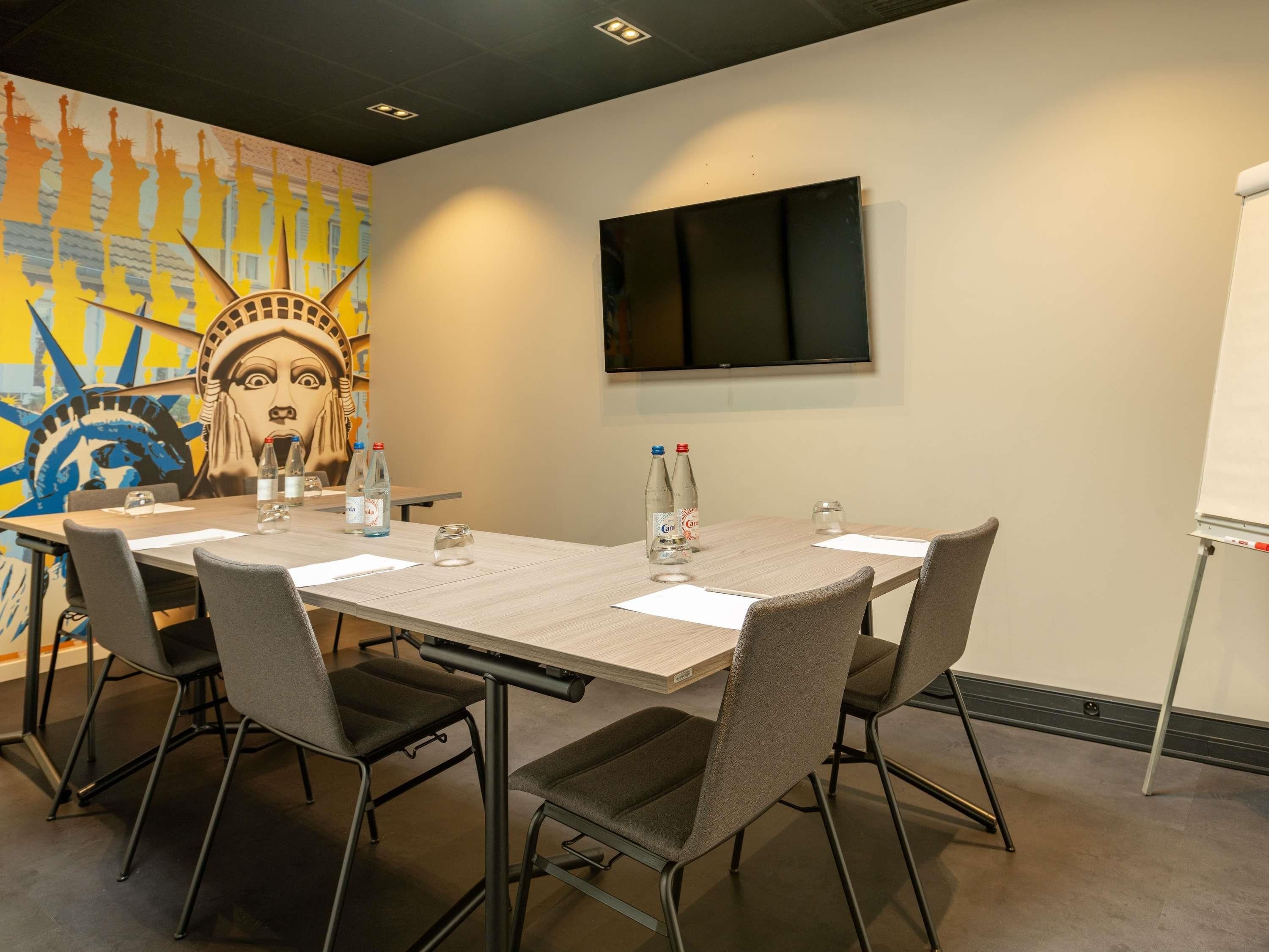 A conference room with a long table, grey chairs, a TV mounted on the wall, and a mural of the Statue of Liberty.