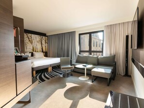 A hotel room with a bed, a sofa, a small table, a TV, and a window with curtains. - Novotel Suites Colmar Centre (Colmar)