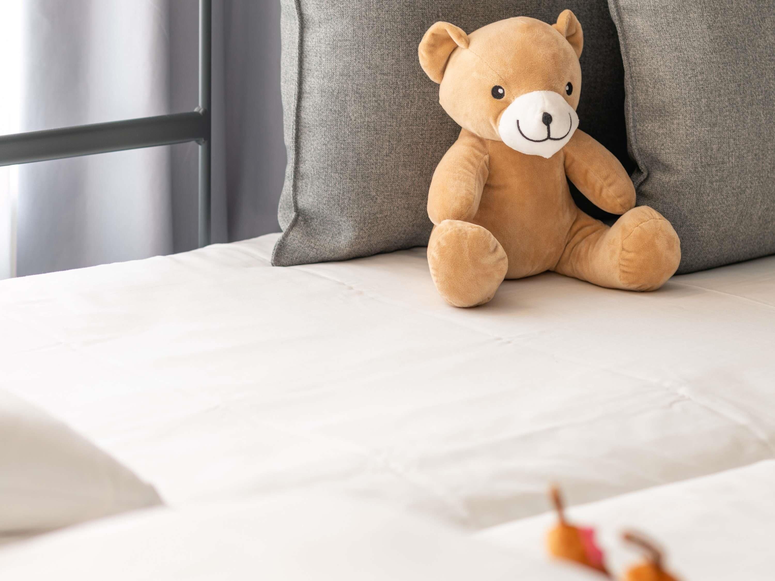 A plush teddy bear sitting on a bed with a grey pillow and a white sheet.