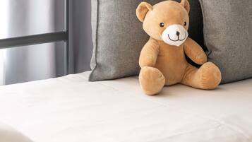 A plush teddy bear sitting on a bed with a grey pillow and a white sheet.