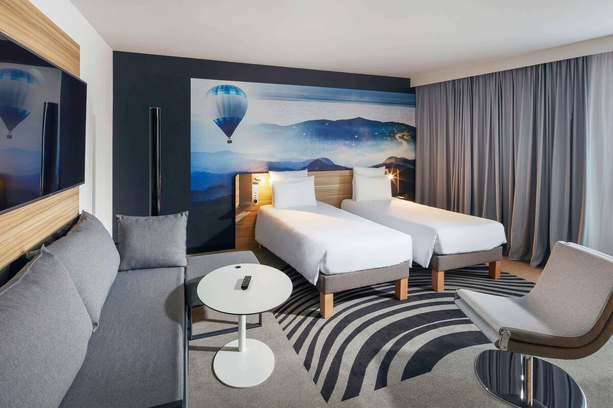 Quarto de hotel com duas camas, um sofá, uma mesa redonda pequena e uma televisão grande.