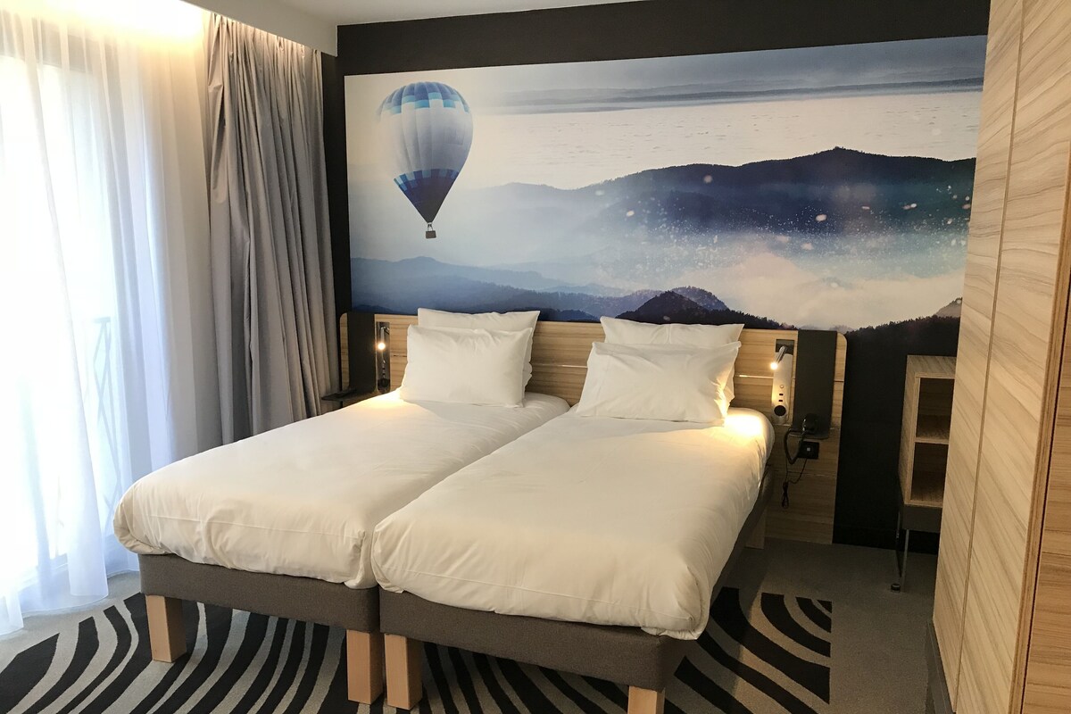 Una cama doble con ropa de cama blanca, una mesita de noche con lámpara y un mural en la pared con un globo aerostático sobre montañas.