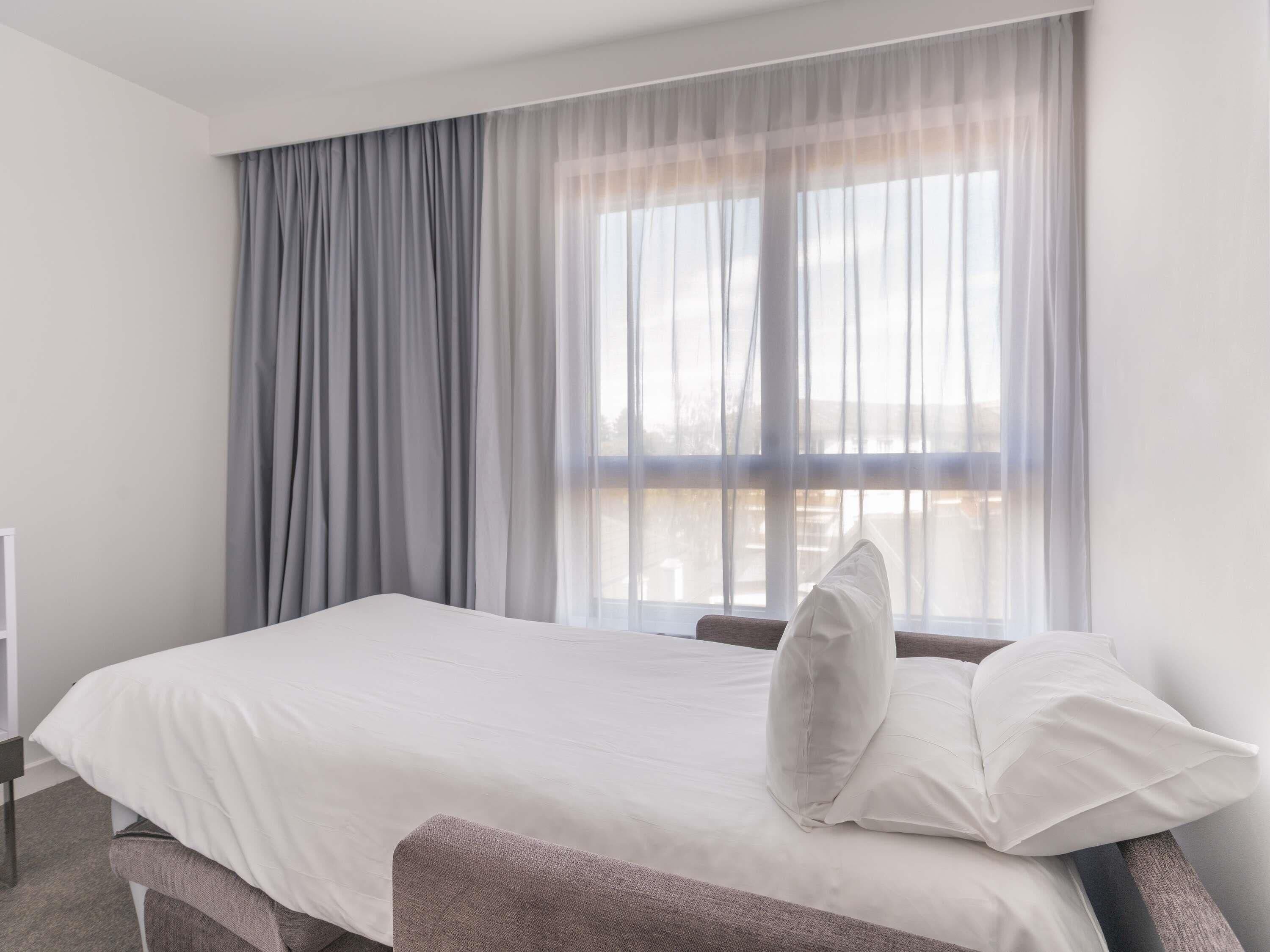 A hotel room with a bed, a window with sheer curtains, and a view of the outside.