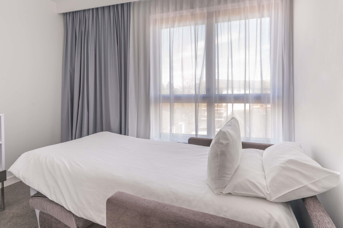 Habitación de hotel con cama, ventana con cortinas translúcidas y vista al exterior.