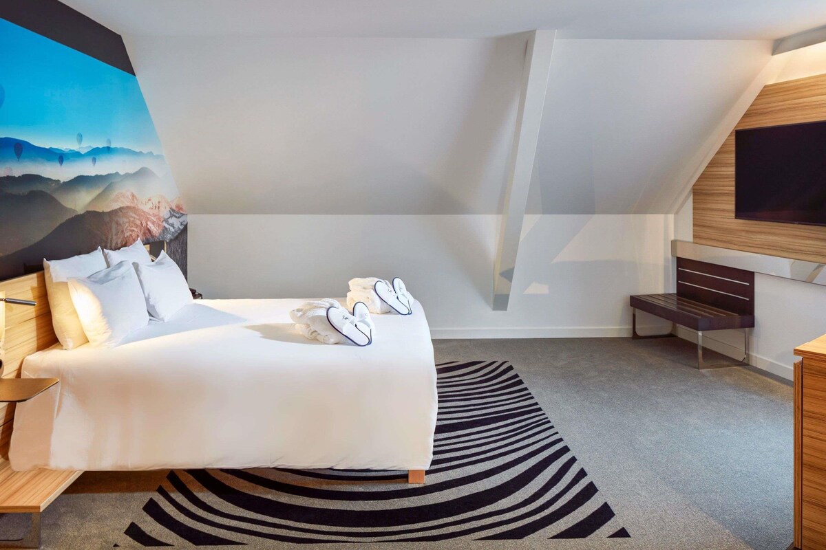 A modern hotel room with a bed, a large painting, a TV, and a bench.
