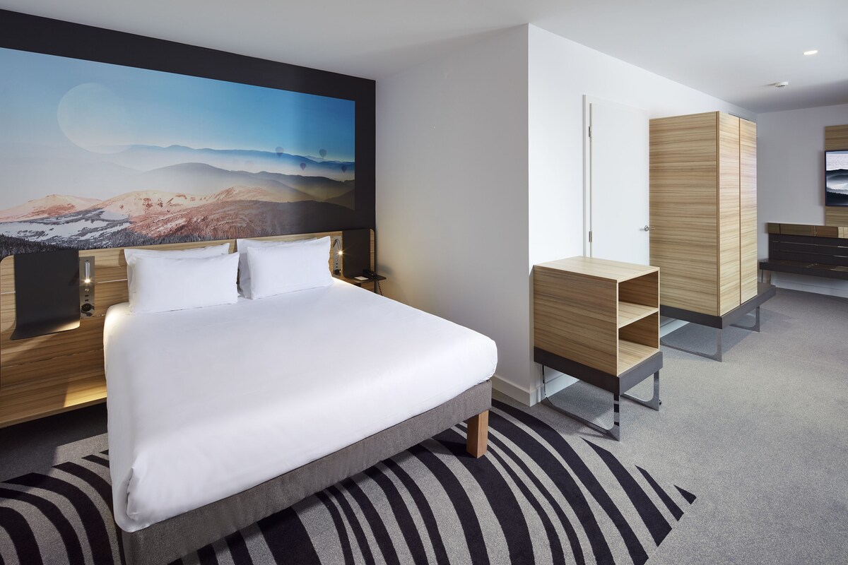 Habitación de hotel moderna con una cama grande, un armario de madera, un televisor de pantalla plana y un mural panorámico en la pared.