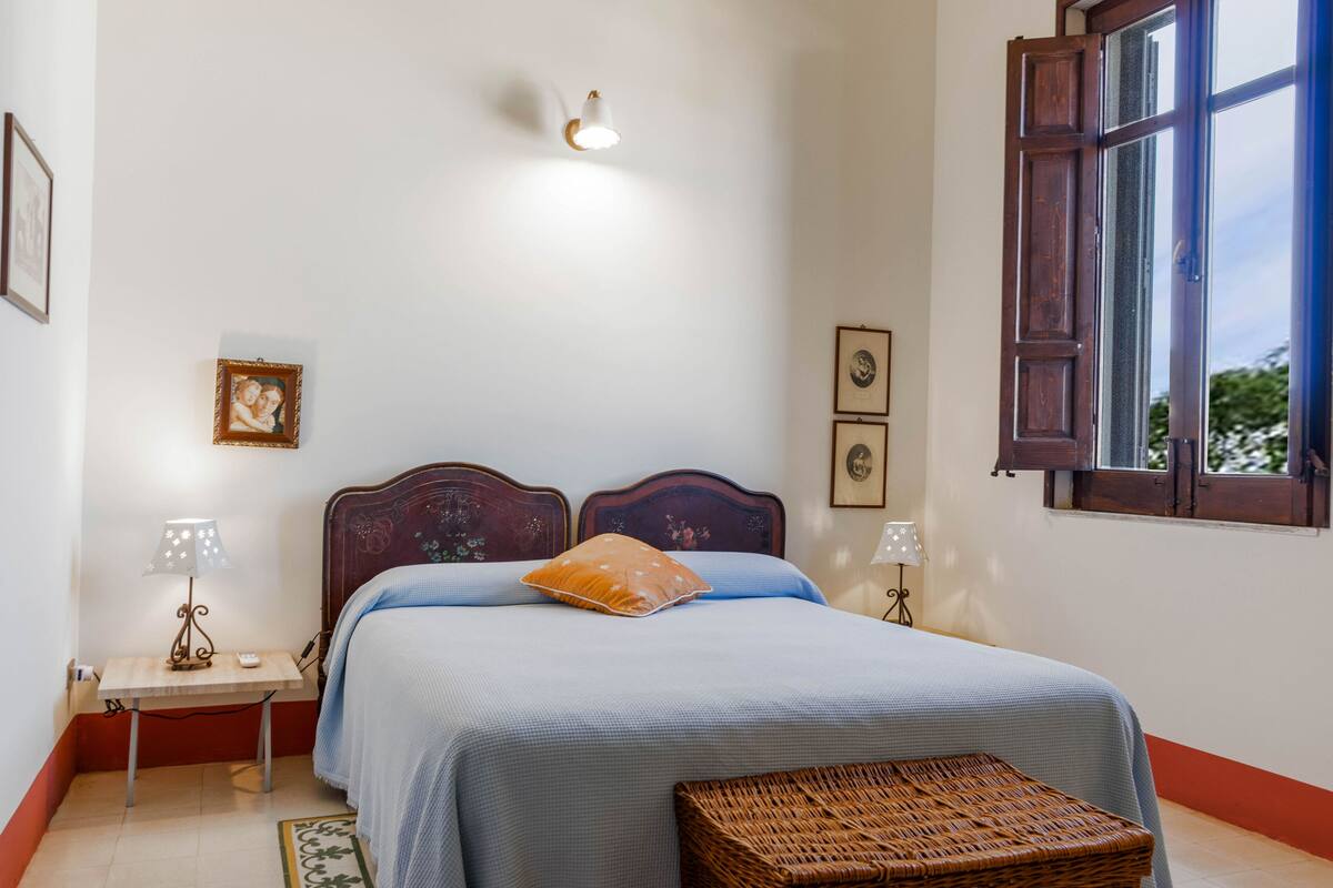 A bedroom with a bed, two bedside tables, a nightstand, a window, and a wall-mounted lamp.