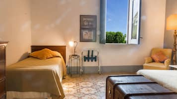 A bedroom with a bed, a chair, a lamp, a suitcase, and a window with a view of greenery.